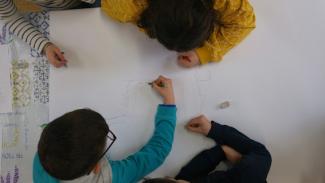 Vue du dessus d'une table sur laquelle trois enfants sont affalés, en train de dessiner sur une feuille de dessin blanche